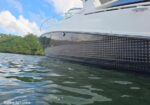 Close-up view of a luxury motorboat's hull at water level, with a dotted pattern along the side and calm water in the foreground, greenery and blue sky in the background