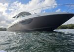 Sleek motor yacht seen from a low side angle on calm water, white hull with dark lower hull and chrome railing, blue sky with scattered clouds above.