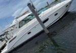 White motor yacht docked at a marina, tied to a piling, with calm water and a blue sky in the background.