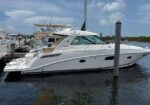 White luxury motor yacht docked at a marina, with other boats and calm water under a blue sky.