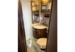 Compact bathroom with a curved wooden vanity, mirrored cabinet, sink, and a toilet; small trash bin on a tiled floor.