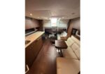 Yacht cabin with a kitchenette on the left, beige sofa and table on the right, and seating toward the back.