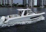 White luxury motor yacht cruising on calm water with a city skyline of high-rise buildings in the background; passengers visible on the deck under a shaded cabin.
