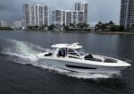 White motor yacht cruising on calm water past a city shoreline with high-rise buildings and palm trees in the background, a person visible at the helm.