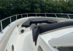 White motorboat bow with black cushioned seating and railing, on calm water with green trees in the background.