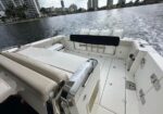 White motorboat deck with beige cushioned seating, chrome railings, and a black backrest; three outboard engines at the stern, calm water and a city skyline in the background.