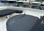 White boat deck with dark gray cushioned seating, railing, and a hatch, with several moored yachts visible in a marina background