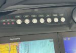 Close-up of a boat cockpit control panel with labeled switches (Bilge, Horn, Mode, Shade, Vent, Washer, Wiper) and red indicator lights; Raymarine display below shows navigation charts.