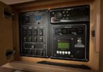 Electrical control panel mounted in a wooden cabinet, featuring battery switch bank, voltmeter, and labeled toggles on the left, plus a stereo and generator monitor on the right.