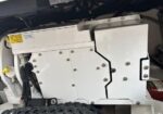 Close-up of a white metal drive housing on heavy equipment, showing bolts, cables, a warning label, and a rugged tire in the foreground.