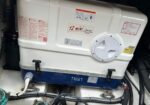 White rectangular tank with a circular hatch, mounted on a blue base, in a mechanical compartment with warning labels and cables around it.