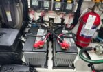 Two gray marine batteries connected in parallel with red terminal clamps in a boat’s electrical compartment, beside a red fire extinguisher.