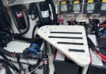 Close-up of a vehicle’s electrical compartment: a white, slotted metal cover over a battery, with red and black cables, hoses, and fuses in the background.