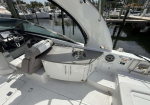 Yacht cockpit area with a gray kitchenette and sink, cup holder, helm chair, open canopy, marina with boats in background.