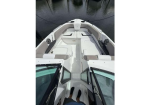 Top-down view of a white motorboat's bow seating area: cushioned benches along the sides with a center walkway, open cockpit at the bottom, two people standing at the stern wearing dark socks, water visible surrounding the boat.