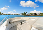 Deck of a white yacht with beige cushions and a small table, overlooking blue water and distant shoreline houses on a sunny day.