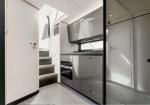 Compact, modern kitchenette with glossy grey cabinets, oven, and stove along a narrow passage, stairs on the left, and white cabinetry overhead.