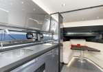 Interior of a modern luxury boat: a sleek galley with a sink and gray cabinets, and a white cushioned seating area with a black table nearby.