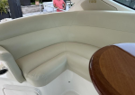 Cream-colored curved boat seating with stitched details and a polished wood table on a white deck; man in a pink shirt stands in the background outdoors.