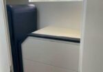 Corner office nook with a light beige desk surface and a dark blue padded panel on the left, against light walls.