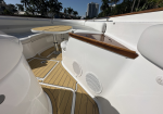 White yacht deck with a round wood-table and curved white seating, teak flooring, and a wooden gunwale; palm trees and a high-rise building in the background.
