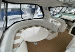 Curved white bench seating around a round white table on a yacht deck, with teak flooring and large cabin windows overlooking other boats in the marina.