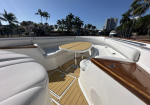 Luxurious white yacht deck with teak floor, curved seating, and a round wooden table under a clear blue sky, overlooking a canal with palm trees and buildings in the background.