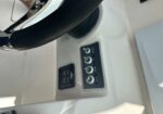 Close-up of a boat helm: black steering wheel with chrome trim and a panel of four circular toggle switches in a dark plate inside a white cockpit area.