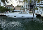White motor yacht moored at a marina beside a concrete dock, with palm trees and a modern building in the background.