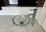 Chrome tall-faucet and clear glass vessel sink on a light countertop, ready for hand washing.