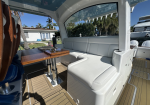 White cushioned L-shaped seating area and a wooden table inside a luxury boat lounge rimmed by a beige deck.