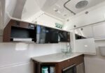 Boat galley/kitchen: wooden cabinets, countertop with glass sink, mounted TV and microwave, ocean-white walls and striped boat flooring.