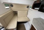 Cream-colored L-shaped seating area in a compact boat cabin with beige cushions, wood paneling, and a small marble-like table surface nearby, plus a wall-mounted TV and two porthole windows.