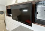 Flat-screen Jensen TV mounted on a wood panel in a boat’s salon, with beige seating nearby and a windowed cabinet to the right.