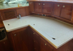Yacht galley with brown wood cabinetry and a speckled beige countertop, sink faucet on left and cabinet doors above and below. Egress to water visible through window.