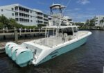 White center-console boat with four turquoise outboard engines docked at a marina, modern buildings in the background.