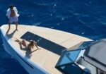 Man standing shirtless in striped swim trunks and a woman in a bikini sunbathe on a yacht deck over deep blue ocean.