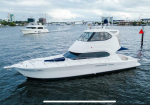 White motor yacht anchored on calm water with a city skyline and other boats in the background.