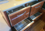 Open kitchen drawer unit showing multiple wooden drawers with metal wire racks and a beige countertop above.