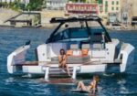White luxury motor yacht anchored near a coastal town; people sunbathe on the rear deck and swim in the water nearby.