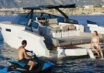 White luxury yacht in blue water with a man on a blue jet ski near the stern and a woman in a white bikini lounging on the swim platform.