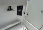 Boat battery control panel with a red circular switch labeled Battery, mounted in a white storage wall; chrome grab bar with black cords to the left. A white door with hinge and latch is on the right.