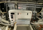 Industrial washing machine in a cluttered mechanical room with pipes and cables overhead and around it.