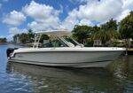 White motorboat with a T-top, docked on a calm canal near palm-lined waterfront under a blue sky with fluffy clouds.