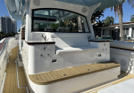 Rear cockpit of a white luxury motorboat with a cushioned bench, wood trim, and built-in storage drawers on teak decking, palm trees and buildings in the background.
