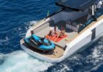 Two people lounge on an orange mat on the rear deck of a luxury boat, while a blue jet ski sits beside them on the deck.