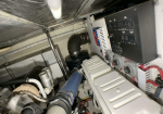 Cramped engine room with a black control panel on the right, large blue hose, and tangled pipes and wiring surrounding heavy machinery.