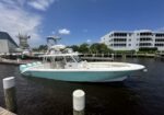 White and turquoise motorboat docked at a marina with a multi-story residential building and blue sky in the background