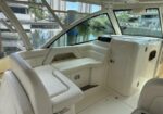 White boat cabin seating area with curved bench seating and cream upholstery, glass windows, and a helm console nearby, overlooking a marina.