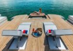 Woman lounging on the edge of a wooden yacht deck, looking out at turquoise sea.